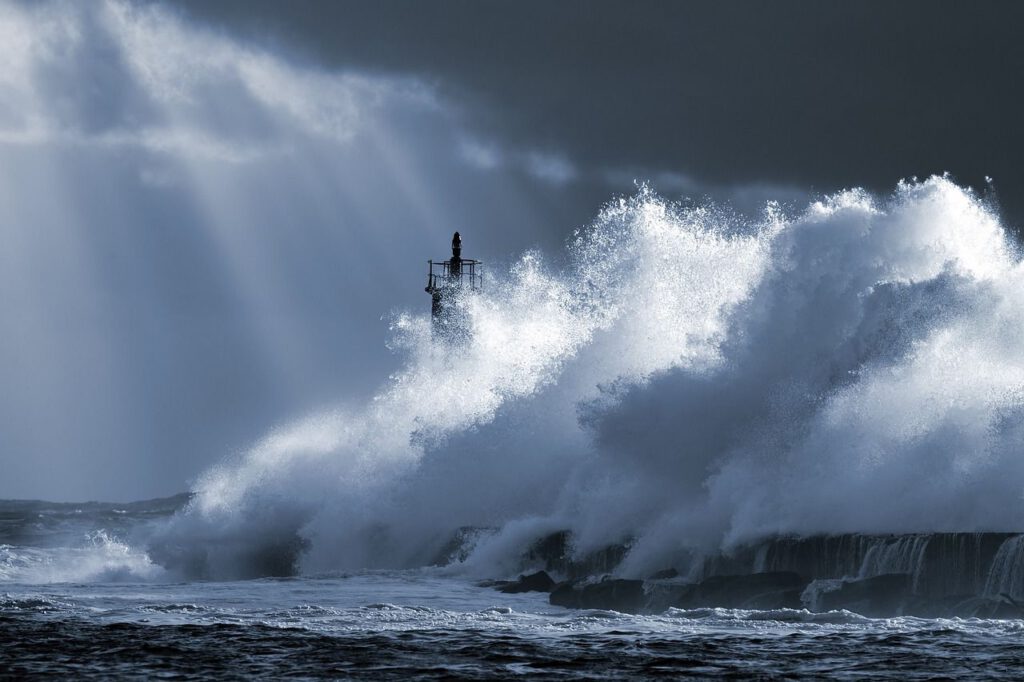 Das Bild zeigt eine stürmische Szene: Ein Hafenkai wird von heftiger Gischt und rauer See regelrecht gepeinigt; die Spitze des Leuchtturms ist bei dem Sturm nur noch knapp zu erkennen. Aus dem drohenden, dunkleren Himmel erstrahlen helle Lichtstreifen.