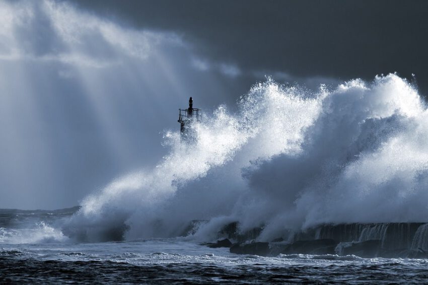 Das Bild zeigt eine stürmische Szene: Ein Hafenkai wird von heftiger Gischt und rauer See regelrecht gepeinigt; die Spitze des Leuchtturms ist bei dem Sturm nur noch knapp zu erkennen. Aus dem drohenden, dunkleren Himmel erstrahlen helle Lichtstreifen.