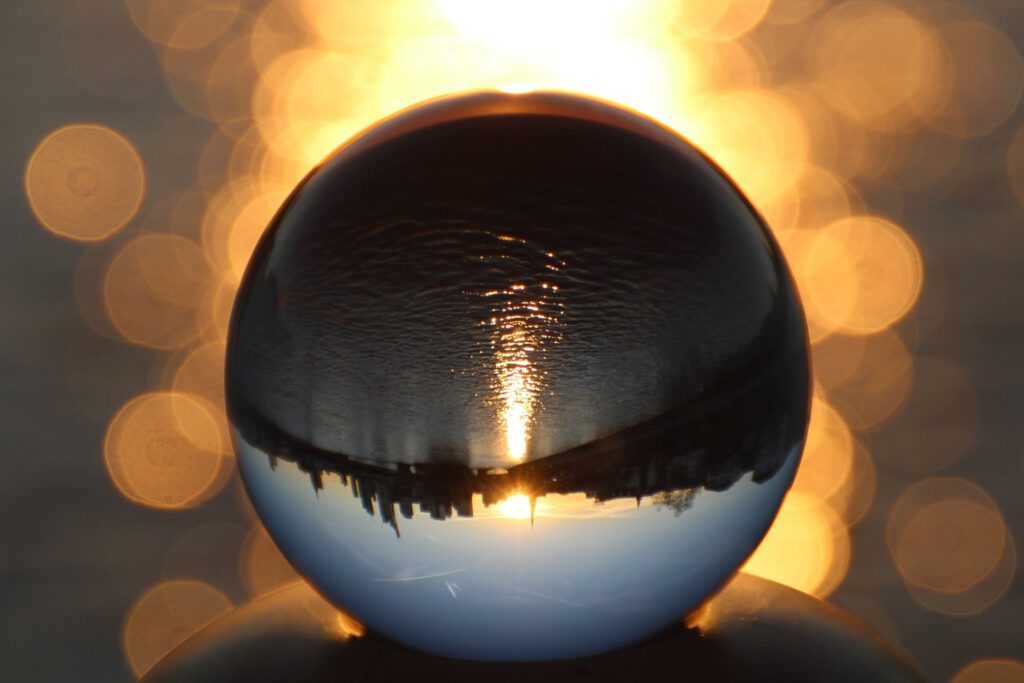 Das Bild zeigt eine Kugel auf leicht schimmerndem, Kupfer-orangenem Hintergrund. Auf der Kugel reflektiert sich das umgekehrte Bild der Stadt Frankfurt und des Flusses Main, bei aufgehender Sonne.