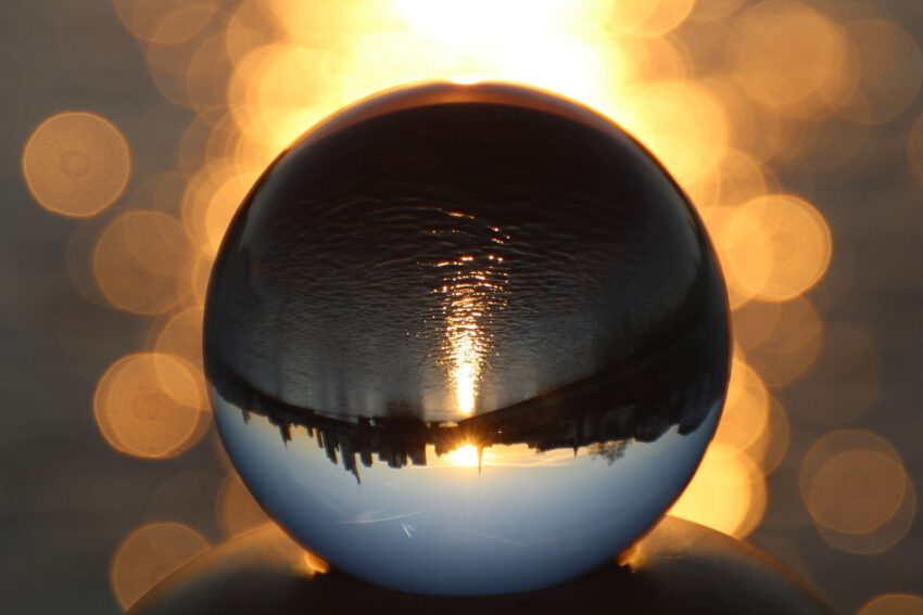 Das Bild zeigt eine Kugel auf leicht schimmerndem, Kupfer-orangenem Hintergrund. Auf der Kugel reflektiert sich das umgekehrte Bild der Stadt Frankfurt und des Flusses Main, bei aufgehender Sonne.
