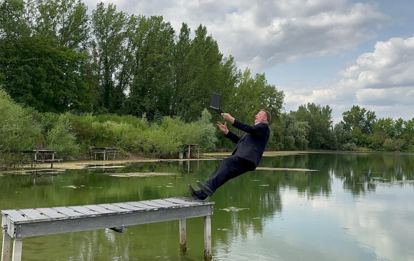Ein Geschäftsmann im Anzug (das ist auch Clark Ahten persönlich) fällt am Ende eines Pontons rückwärts in den See, Laptop in der Hand.
